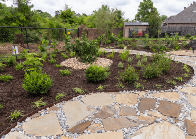 Finished landscape project. Flagstone area with creek rock, metal edging around a flower bed with brown mulch. Bushes and small ground cover plants are in a circle around a big bush.