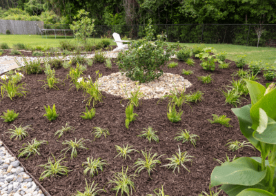 Finished landscape project. Flagstone area with creek rock, metal edging around a flower bed with brown mulch. Bushes and small ground cover plants are in a circle around a big bush.