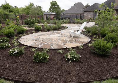 A circular flower bed with brown mulch around a flagstone fire pit area. Small, green shrubs are placed around it.
