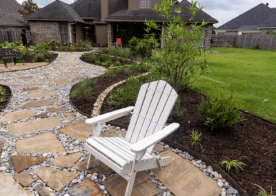 A white chair sitting on a flagstone pathway. Behind it is a new flower bed with brown mulch and green plants.